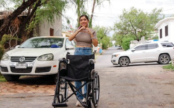 Perla Cavazos durante una visita a una familia de Ciénega de Flores, con una silla de ruedas y un gesto de corazón con las manos