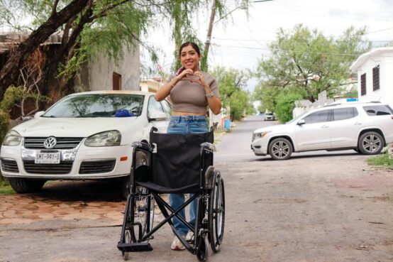 Perla Cavazos durante una visita a una familia de Ciénega de Flores, con una silla de ruedas y un gesto de corazón con las manos