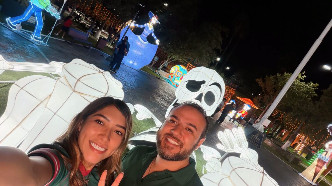 Figuras iluminadas de Día de Muertos en la Plaza Principal de Ciénega de Flores, con dos personas en primer plano.