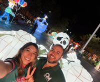 Figuras iluminadas de Día de Muertos en la Plaza Principal de Ciénega de Flores, con dos personas en primer plano.