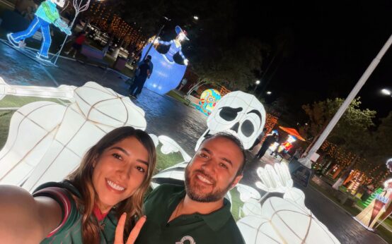 Figuras iluminadas de Día de Muertos en la Plaza Principal de Ciénega de Flores, con dos personas en primer plano.