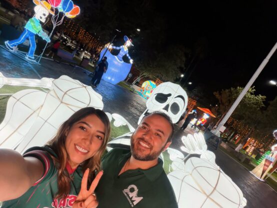 Figuras iluminadas de Día de Muertos en la Plaza Principal de Ciénega de Flores, con dos personas en primer plano.
